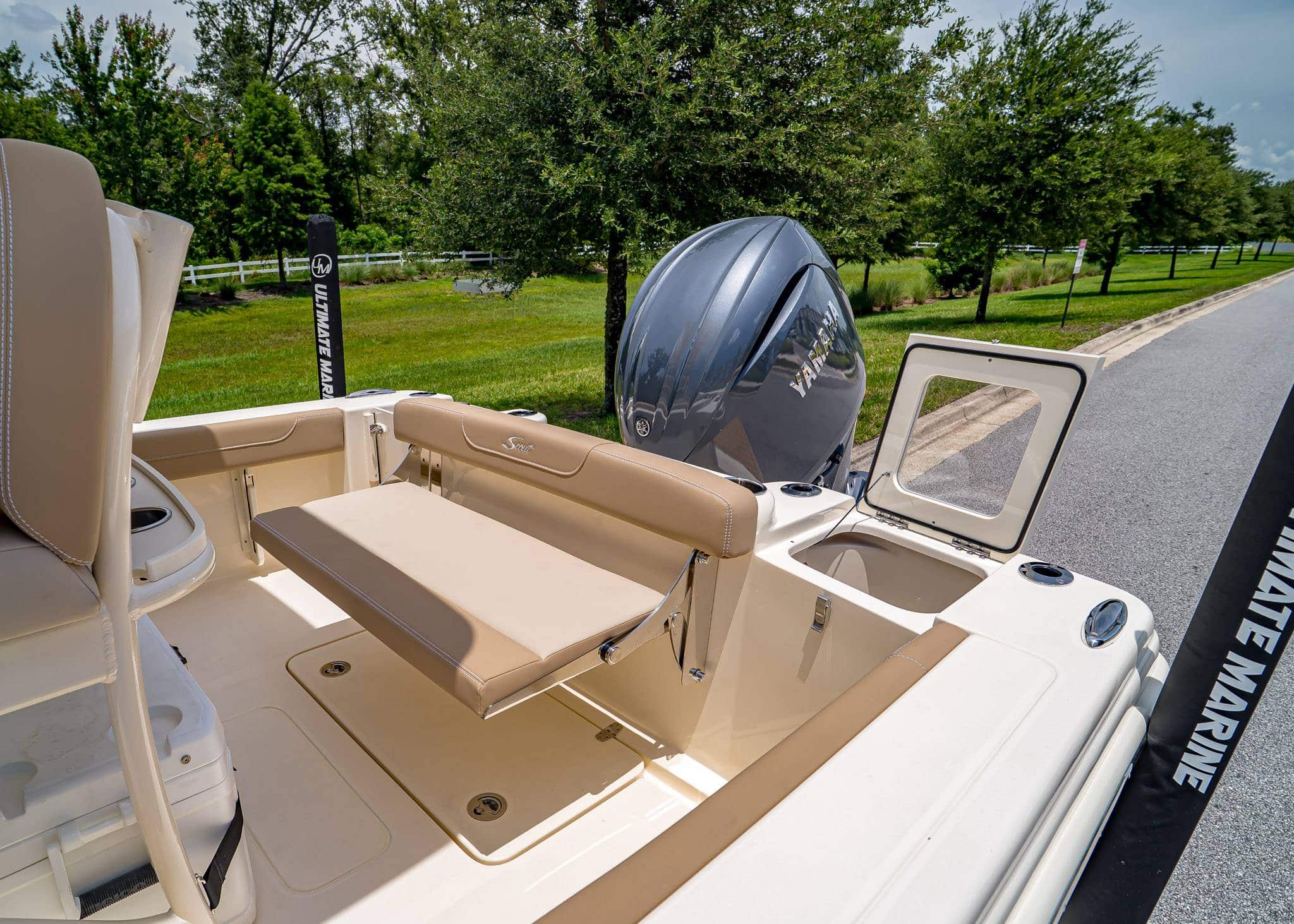 A beige and white Scout 215 boat with a fold-down bench seat, outboard motor, and open storage compartment, parked on a paved road beside grass and trees.