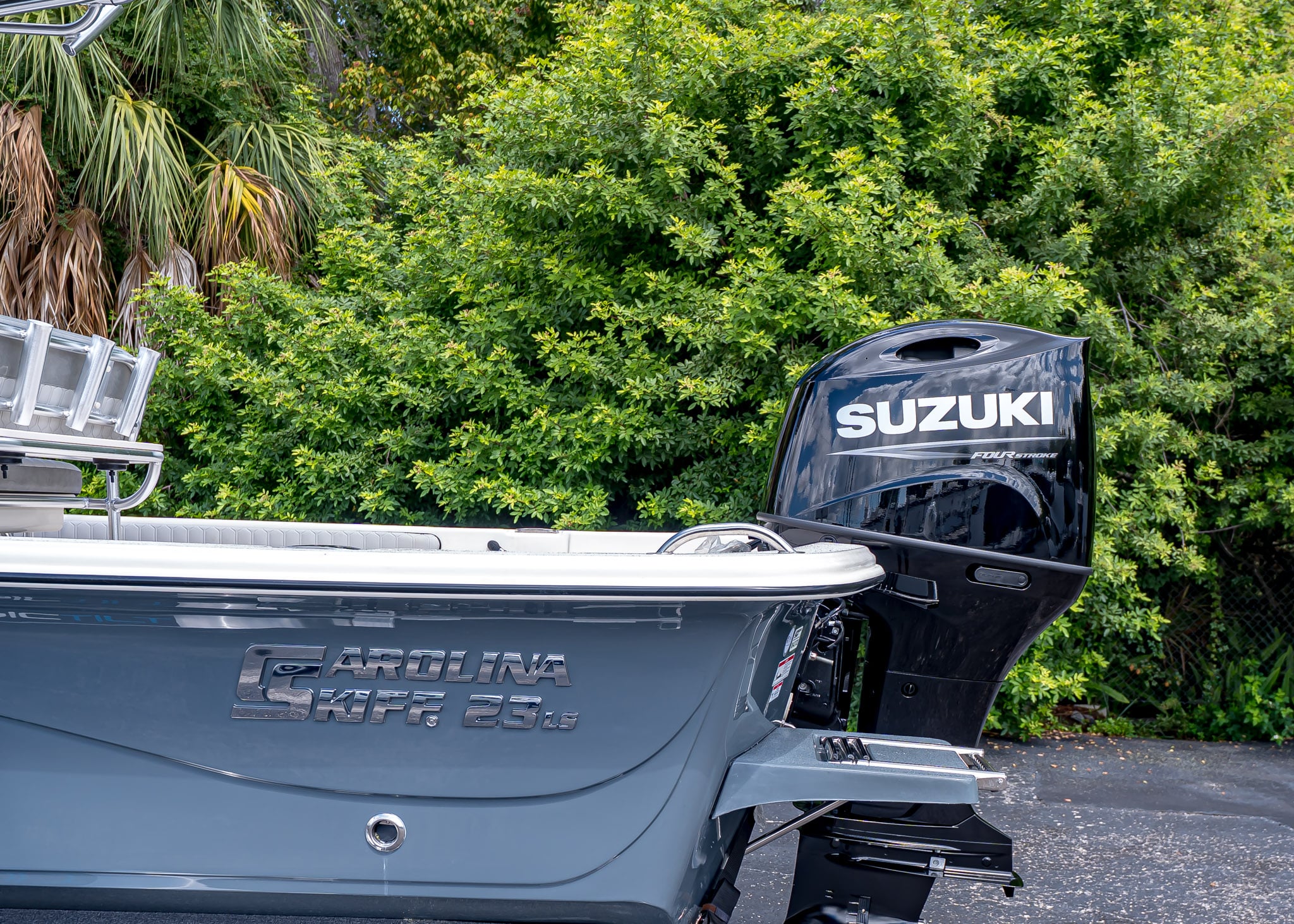 A gray Carolina Skiff 23 LS—often called the best entry boat—rests on asphalt, featuring a black Suzuki outboard motor and framed by lush green foliage in the background.